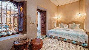 Een slaapkamer met een groot bed, een versierde houten deur en uitzicht door een raam met sierlijk ijzerwerk.