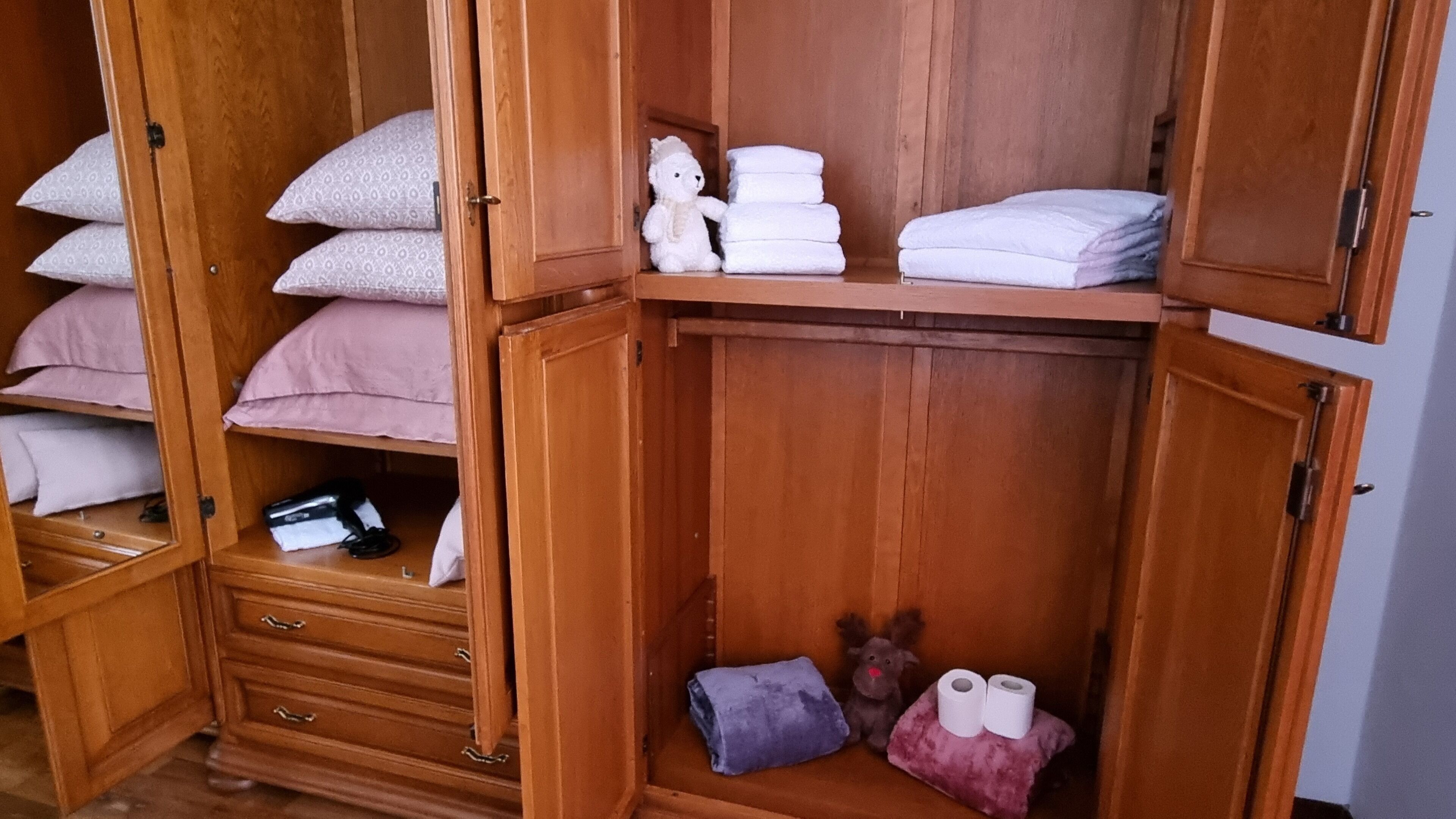 A wooden wardrobe with multiple shelves, some containing folded towels and pillows, and a small shelf with a plush toy and a rolled-up towel.