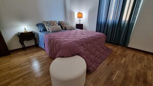 Un dormitori amb un llit, una tauleta de nit, una làmpada i una finestra amb cortines.