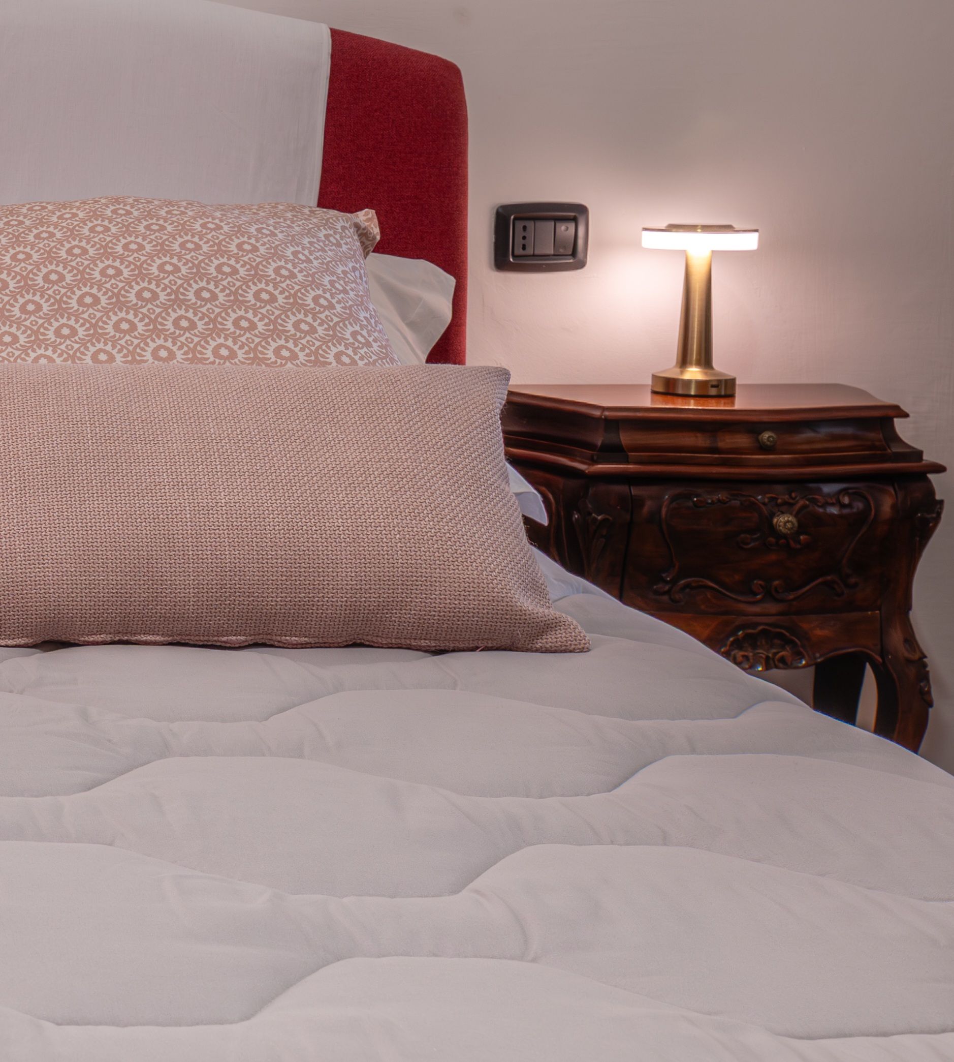 Un lit avec une couette blanche et des oreillers à motifs, une table de chevet avec une lampe et une prise électrique au mur.