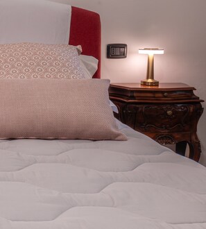Ein Bett mit weißer Daunendecke und gemusterten Kissen, ein Nachttisch mit Lampe sowie eine Wandsteckdose.