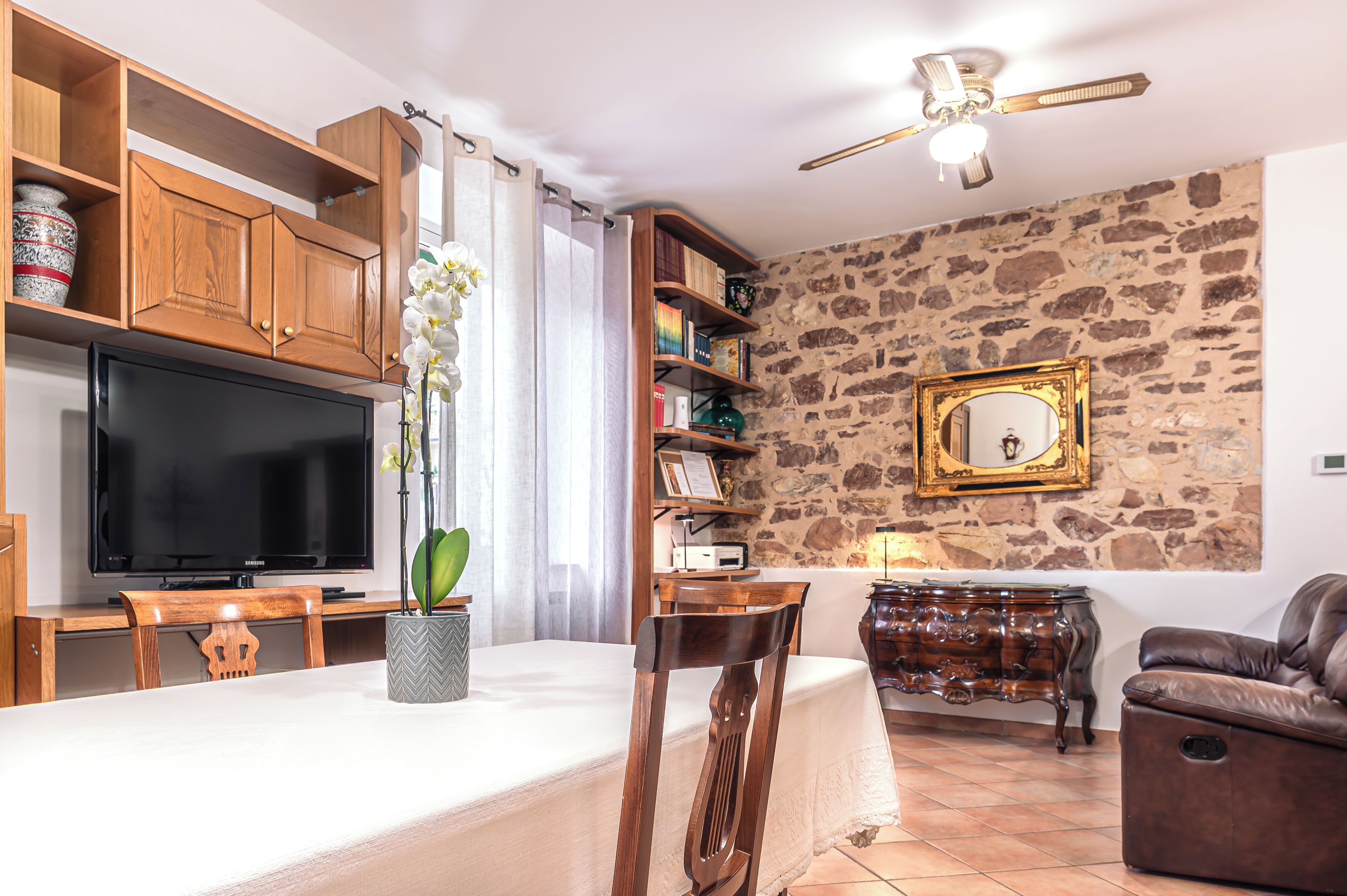 A living room with a television, wooden cabinets, a stone wall, a sofa, and a dining area with chairs and a table.
