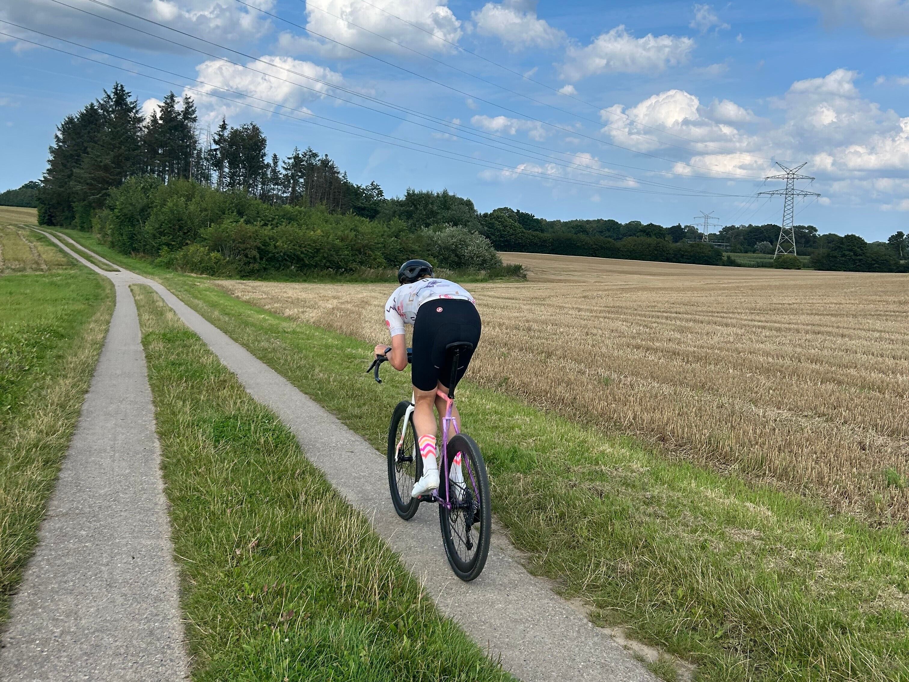 Un cycliste sur une piste asphaltée, dans un cadre rural, avec des champs et des arbres.