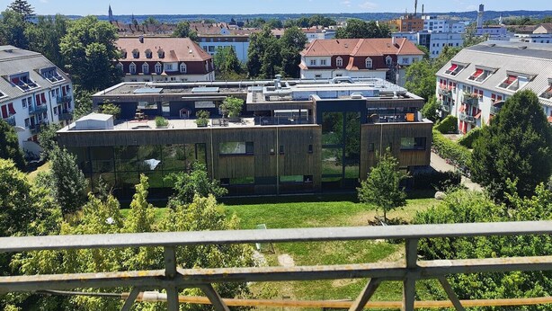 Um edifício moderno com telhado plano, rodeado por árvores e outros edifícios.
