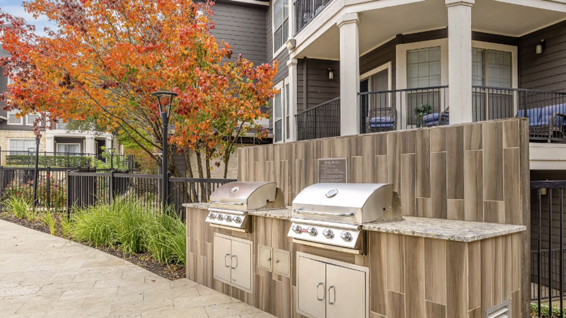 An outdoor kitchen area with a grill and a built-in oven, surrounded by a wooden fence and a tree with autumn leaves.