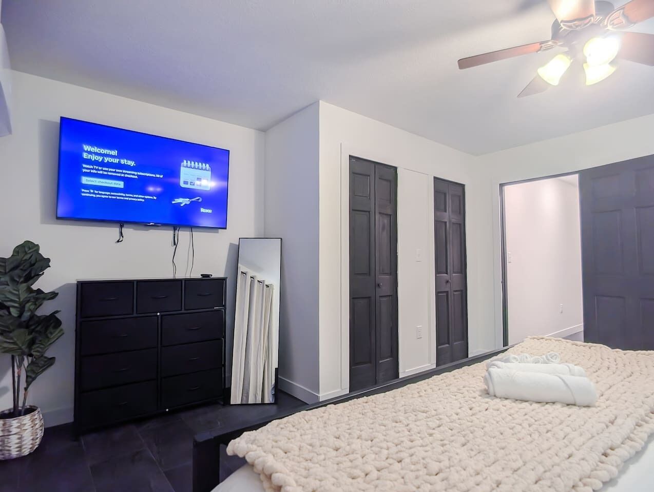 Ein modernes Hotelzimmer mit Flachbildfernseher, einem dunklen Holzschrank, einem großen Spiegel, einem flauschigen weißen Bett und einem Deckenventilator.