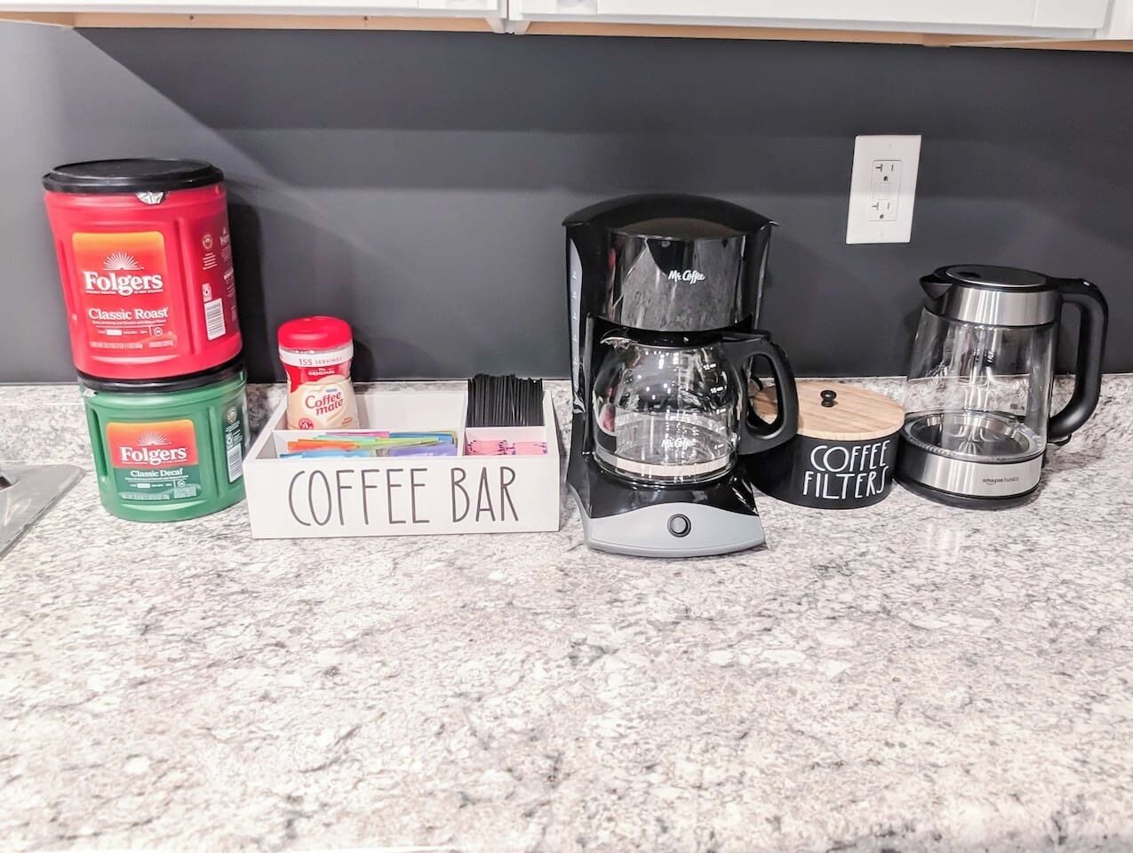 Una encimera de cocina con equipo para preparar café, incluyendo una cafetera, filtros y latas de café Folgers.