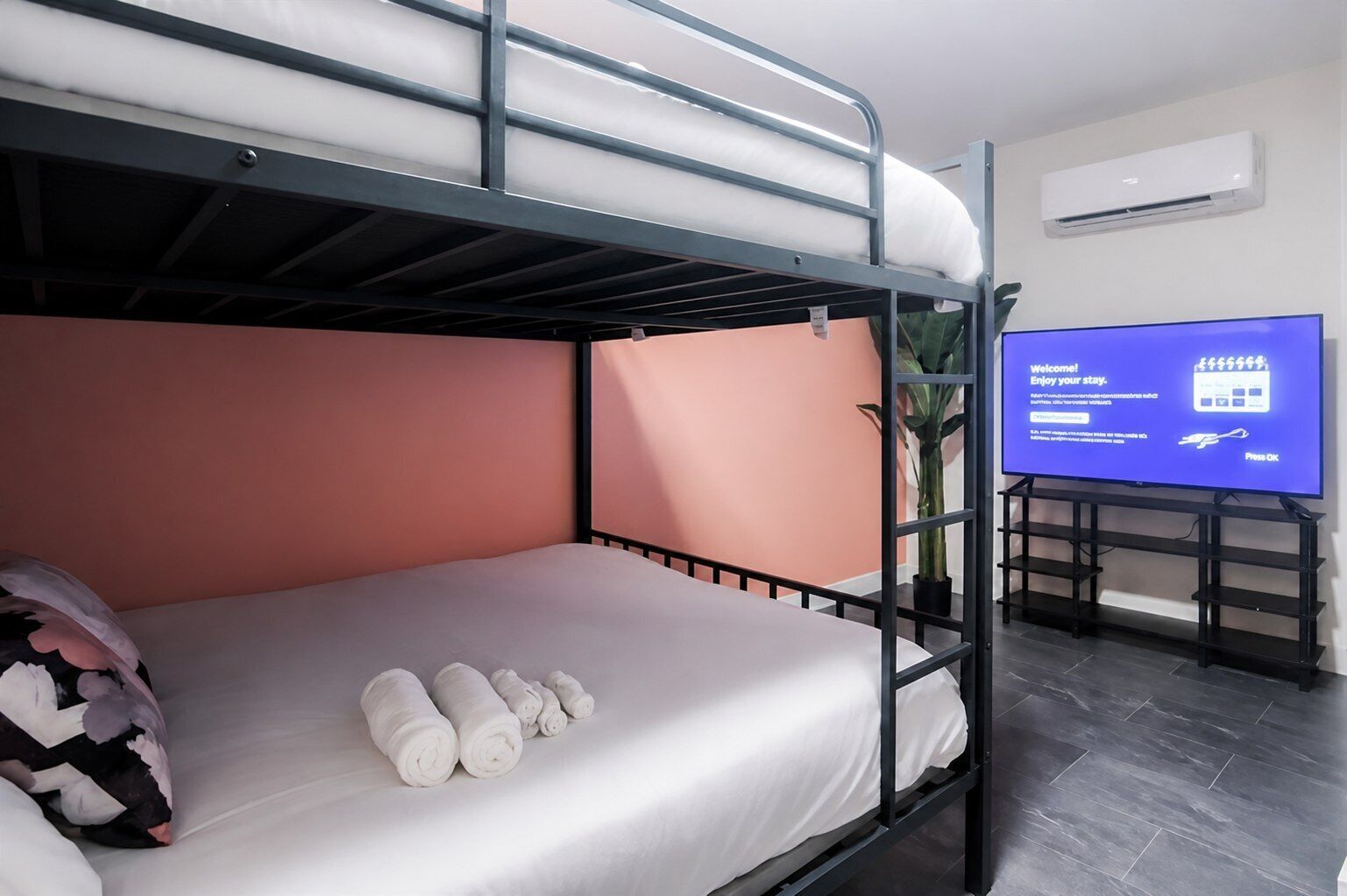Een slaapkamer met stapelbed, een televisie waarop een welkomstboodschap staat, een kamerplant en netjes opgerolde handdoeken op het bed.