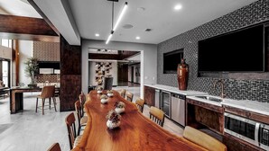 A modern dining area with a long wooden table, chairs, and a kitchen with a tiled backsplash and a large TV. - Landing | Beautiful 2BD, Clubhouse, Rooftop Lounge (San Antonio)