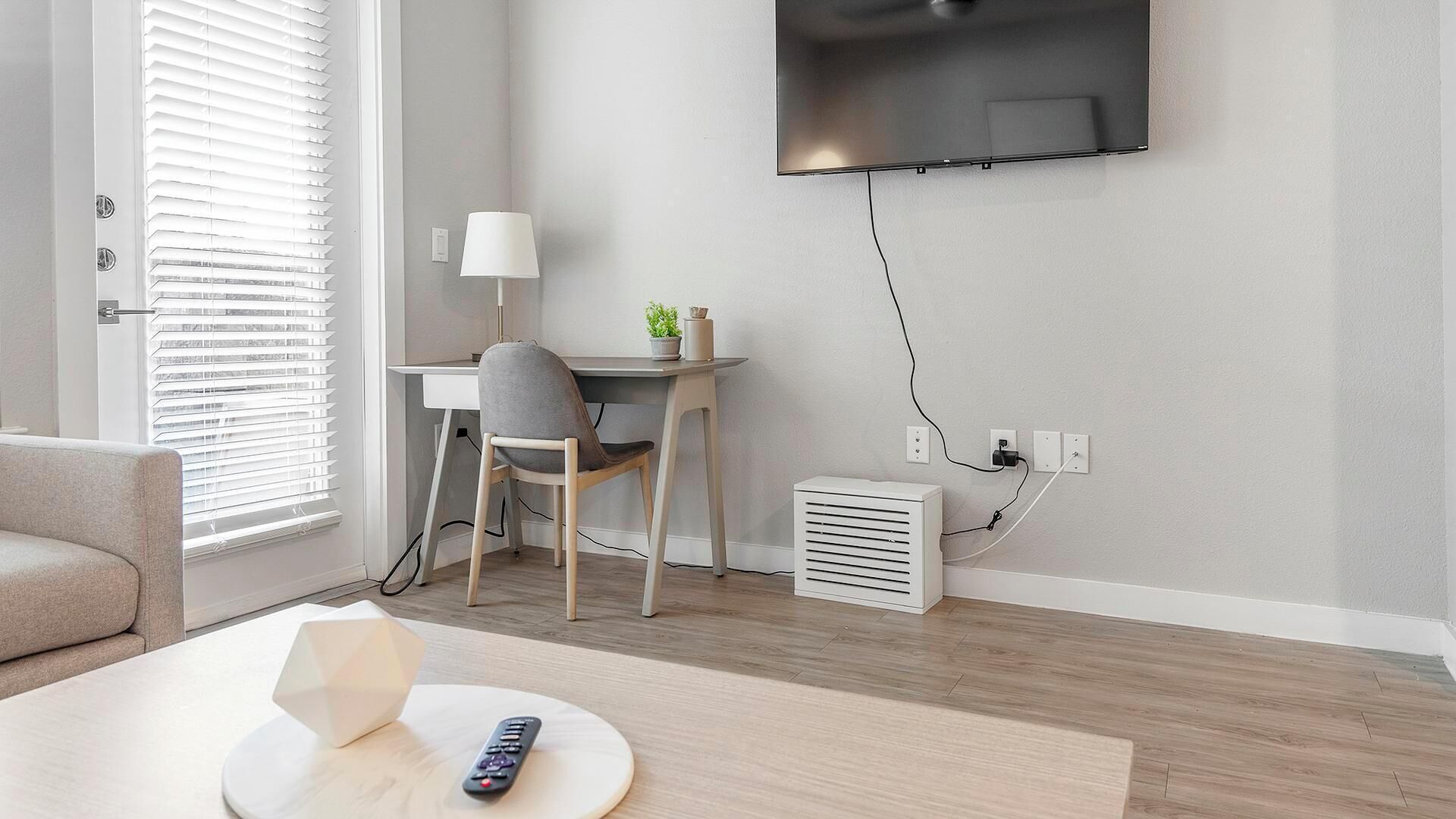 A modern living room with a flat-screen TV mounted on the wall, a desk with a chair, a small table with a lamp, and a heater on the floor.