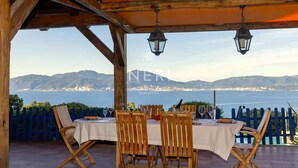 A covered outdoor dining area with wooden chairs and a table set for two, overlooking a scenic coastal view.