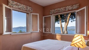 A bedroom with a bed, two windows with curtains, a lamp, and a view of the sea and mountains.