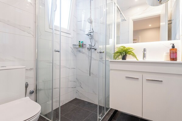Une salle de bain moderne avec une douche à parois de verre, des toilettes blanches, un lavabo avec un miroir et une petite étagère contenant des articles de toilette.