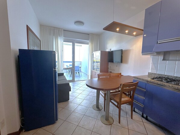 Een compacte keuken met een eethoek, een blauwe koelkast, een flatscreen televisie boven de aanrecht en uitzicht op het balkon door de glazen deur.