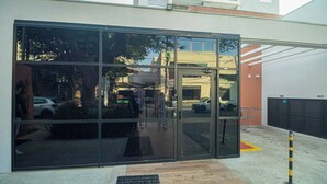 Un edificio con puerta de acceso de vidrio, cerca metálica y zona de estacionamiento con coches.