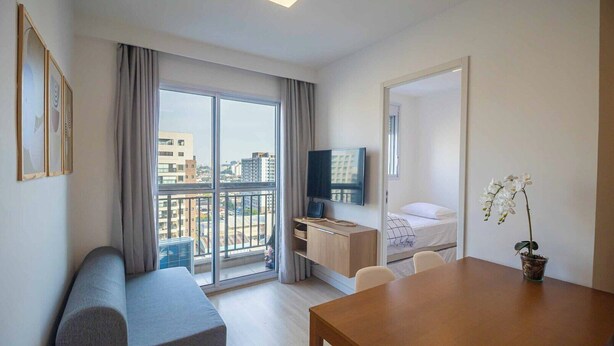 Una habitación de hotel moderna con balcón, una cama, un escritorio, un televisor y una orquídea en maceta.