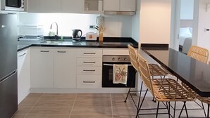 Una cocina moderna con armarios blancos, encimera negra, refrigerador de acero inoxidable, microondas, hervidor y tostadora. Hay una mesa de comedor con sillas y una lámpara colgante.