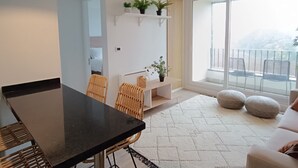 Una sala de estar moderna con comedor, que incluye una mesa negra, sillas de mimbre, un sofá blanco y un amplio ventanal con vistas al exterior.