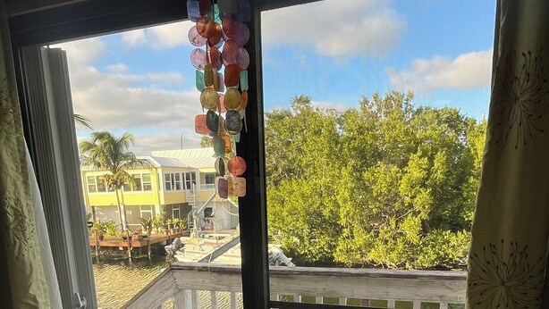 A view from inside a room through a glass door to an outdoor deck with a railing, a palm tree, and a yellow building in the background. - Fisherman's Paradise!  50' Concrete Dock. Space for 2 boats (Big Pine Key)