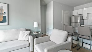 Un salon moderne avec un canapé gris, une petite table d’appoint avec une lampe, et un coin cuisine doté de meubles blancs et d’un réfrigérateur en acier inoxydable.