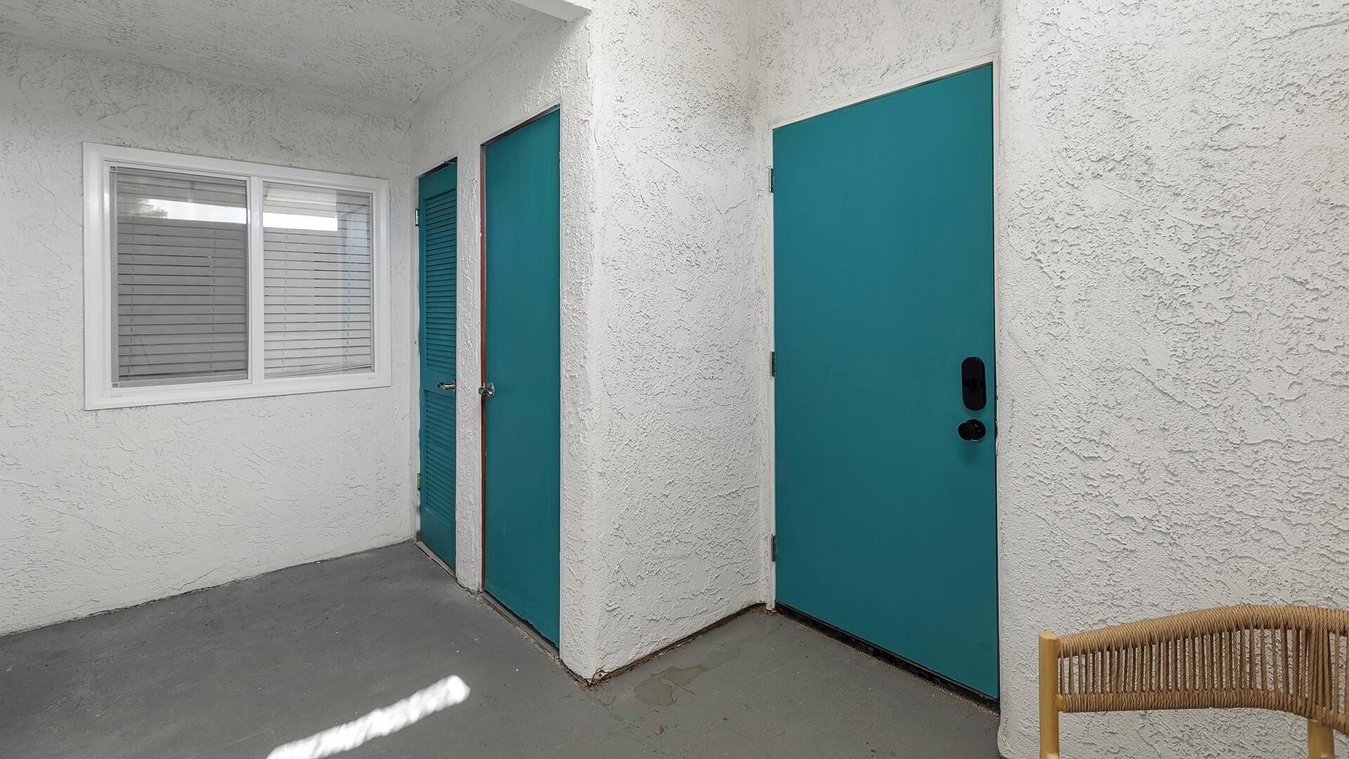 A narrow hallway with white textured walls, a teal door, a white door, and a window with blinds.