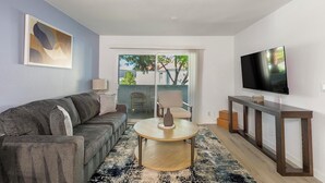 Sala de estar con sofá gris, una mesa redonda, televisor sobre un mueble de madera y vistas al exterior a través de un ventanal.