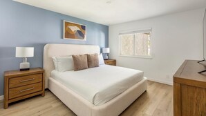 Un dormitorio con una cama, dos mesitas de noche, una lámpara de noche, una ventana y un cuadro en la pared.