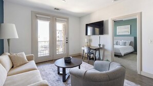 En moderne stue med sofa, lænestol og sofabord. Der er et fjernsyn monteret på væggen, og en balkon med udsigt.