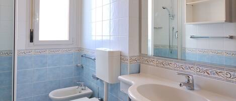 A bathroom with a toilet, bidet, and sink, tiled in blue and white, with a window and a mirror.