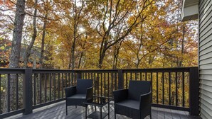 Una terraza de madera con dos sillas de mimbre y una pequeña mesa, con vistas a un bosque de follaje otoñal.