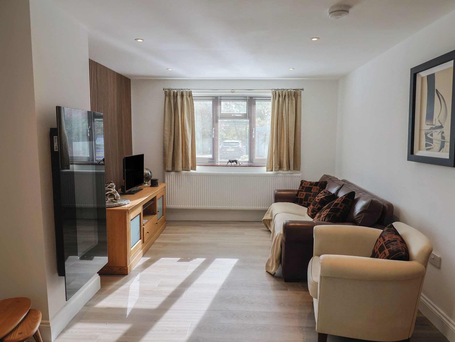 A modern living room with a flat-screen TV, wooden entertainment unit, beige sofa, and a large window with curtains.