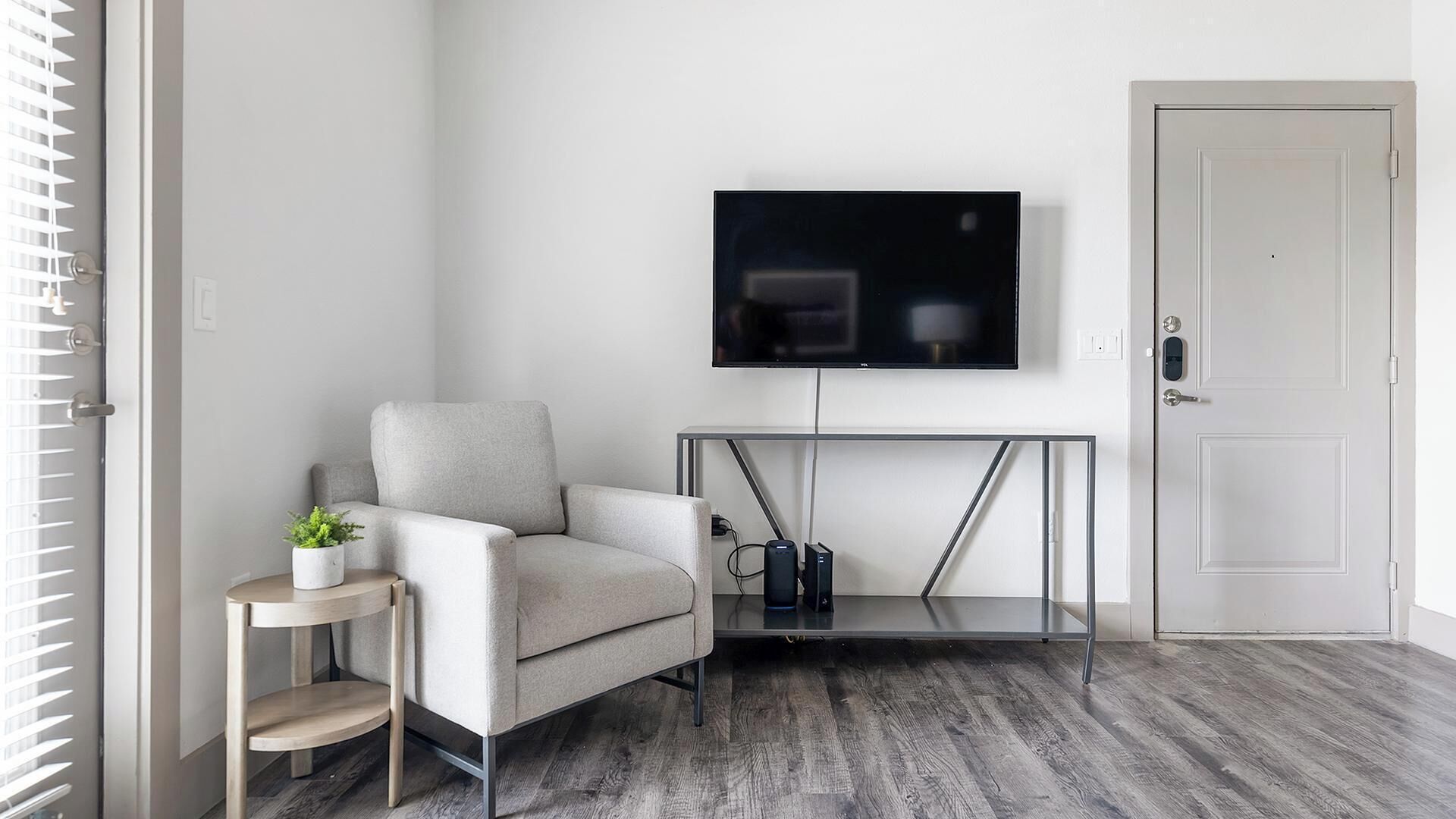 Een moderne woonkamer met een grijze fauteuil, een klein bijzettafeltje met een plant, een aan de muur bevestigde televisie en een deur met een sleutel in het slot.