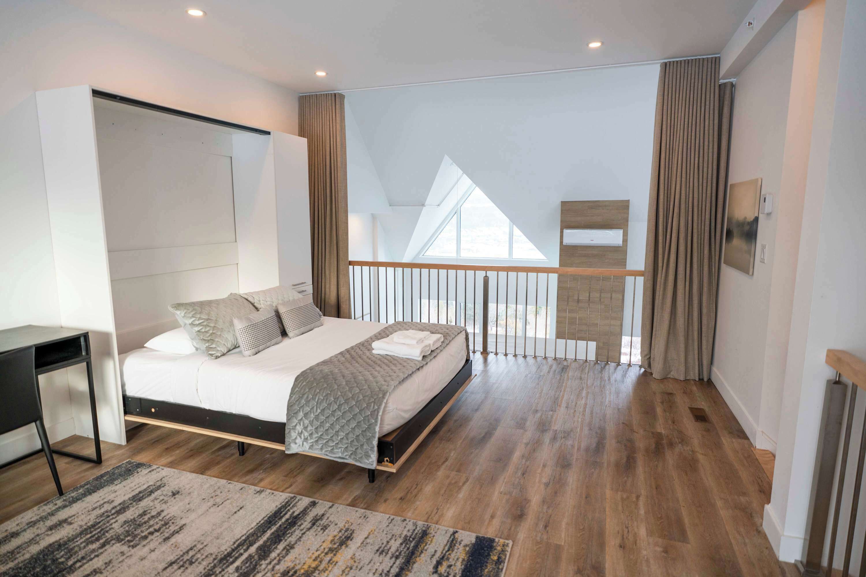 A modern bedroom with a bed, a desk, and a view of the outdoors through a window.