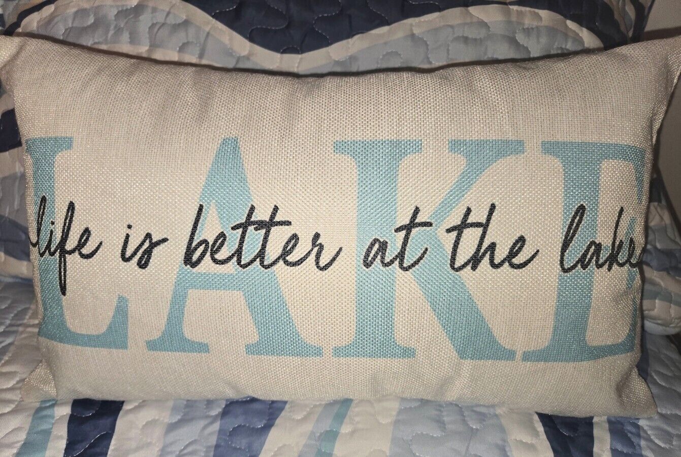 Un coussin orné de la phrase « la vie est plus belle au bord du lac », posé sur un lit avec une literie à rayures.