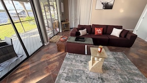 Ein Wohnzimmer mit einer großen Glastür, die auf eine Terrasse führt, ein dunkelbraunes Sofa, ein Glastisch und Blick auf einen Steg und Wasser.