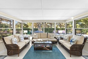 A sunroom with wicker furniture, a glass coffee table, and large windows offering a view of the outdoors. - Coastal Haven Near Beach & Golf (Hilton Head Island)