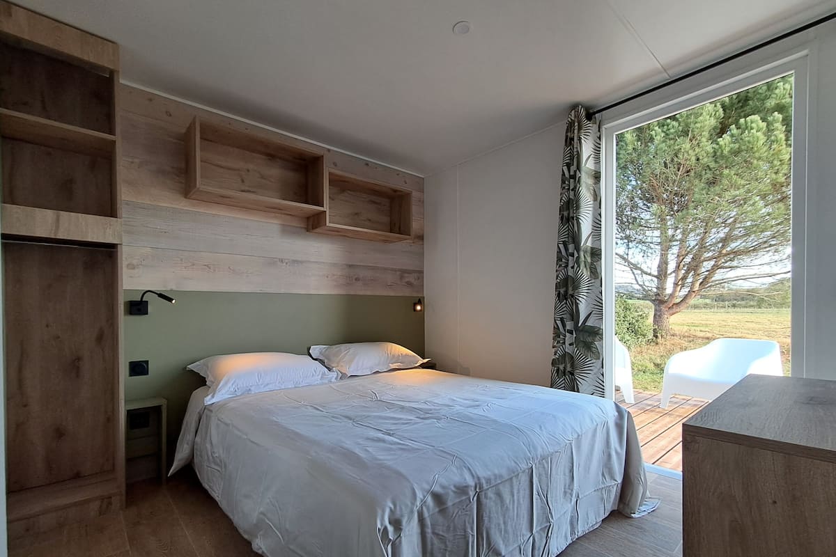 Une chambre à coucher avec un lit, des étagères en bois, une table de chevet et une fenêtre donnant sur des arbres.