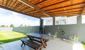 An outdoor patio with a wooden table and benches, a barbecue grill, a striped beach chair, and a potted plant. - House w/ Grill in Front of the Beach Lbra0011 (Ilhéus)