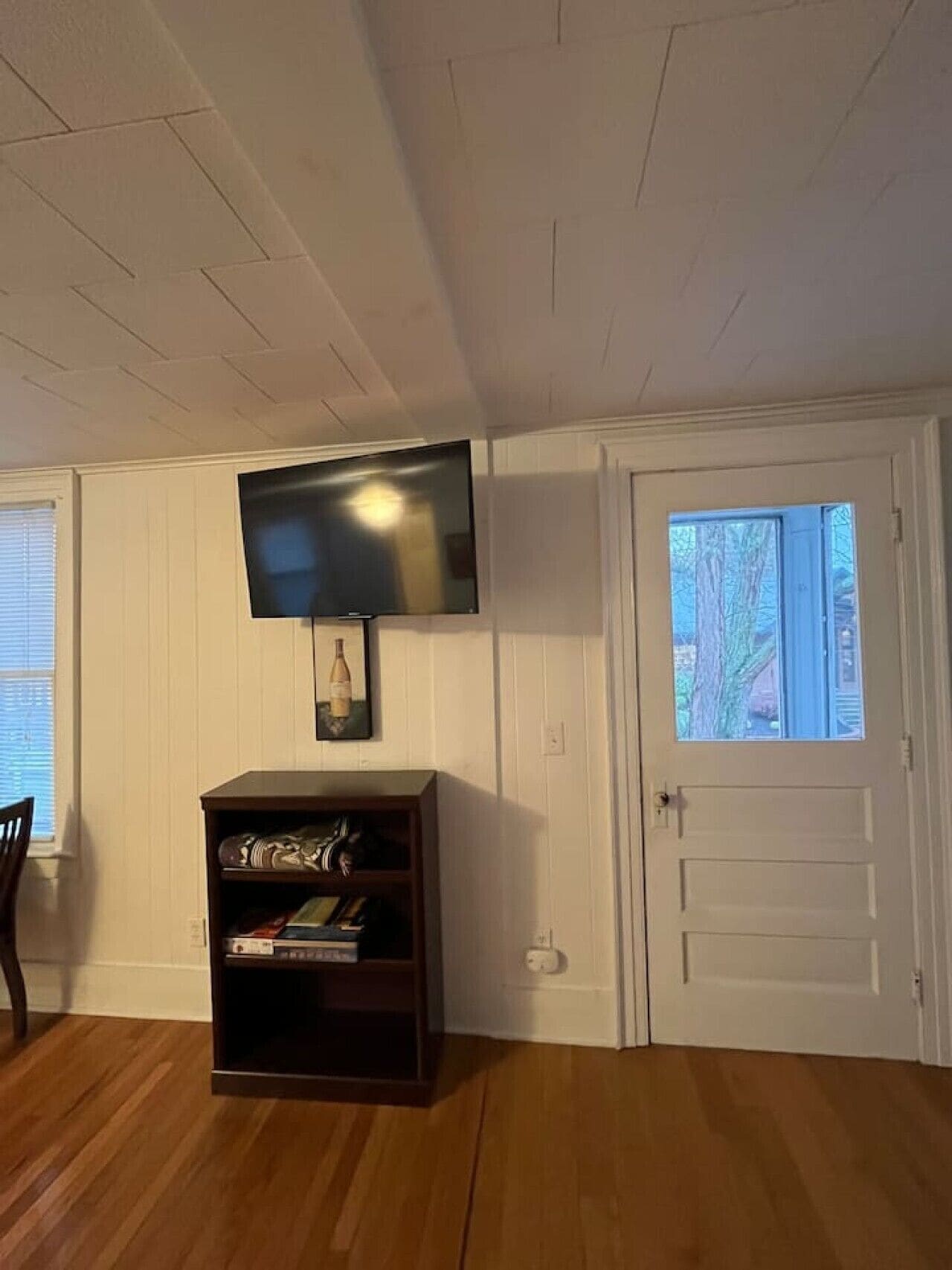 Quarto com televisão de tela plana na parede, uma estante de livros, porta com janela e piso de madeira.