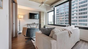 A modern living room with a sofa, a chair, a TV, and a large window with a city view. - Landing | Incredible 1BD, Gym, Game Room (Arlington)