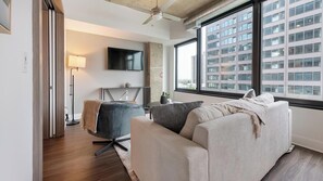 Ein modernes Wohnzimmer mit einer Couch, einem Sessel, einem Fernseher und einem großen Fenster mit Blick auf die Stadt.