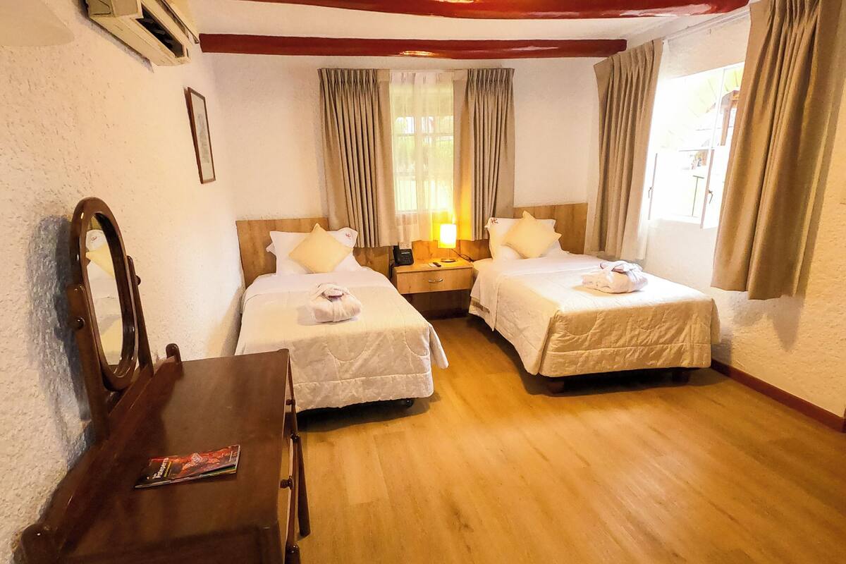 A hotel room with two single beds, a wooden desk, a chair, a mirror, and a window with curtains.
