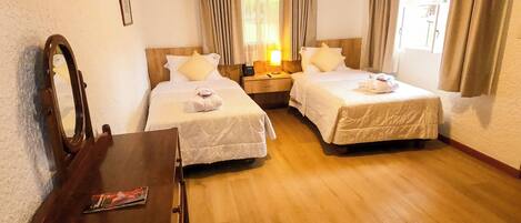 A hotel room with two single beds, a wooden desk, a chair, a mirror, and a window with curtains.