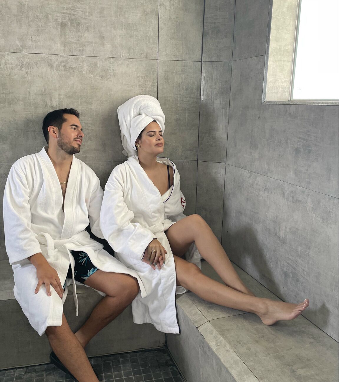 Duas pessoas em roupões brancos sentadas em um banco num quarto com uma janela grande e uma parede revestida de azulejos.
