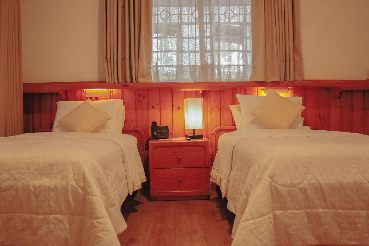 A room with two beds, a nightstand with a lamp, and a window with curtains.