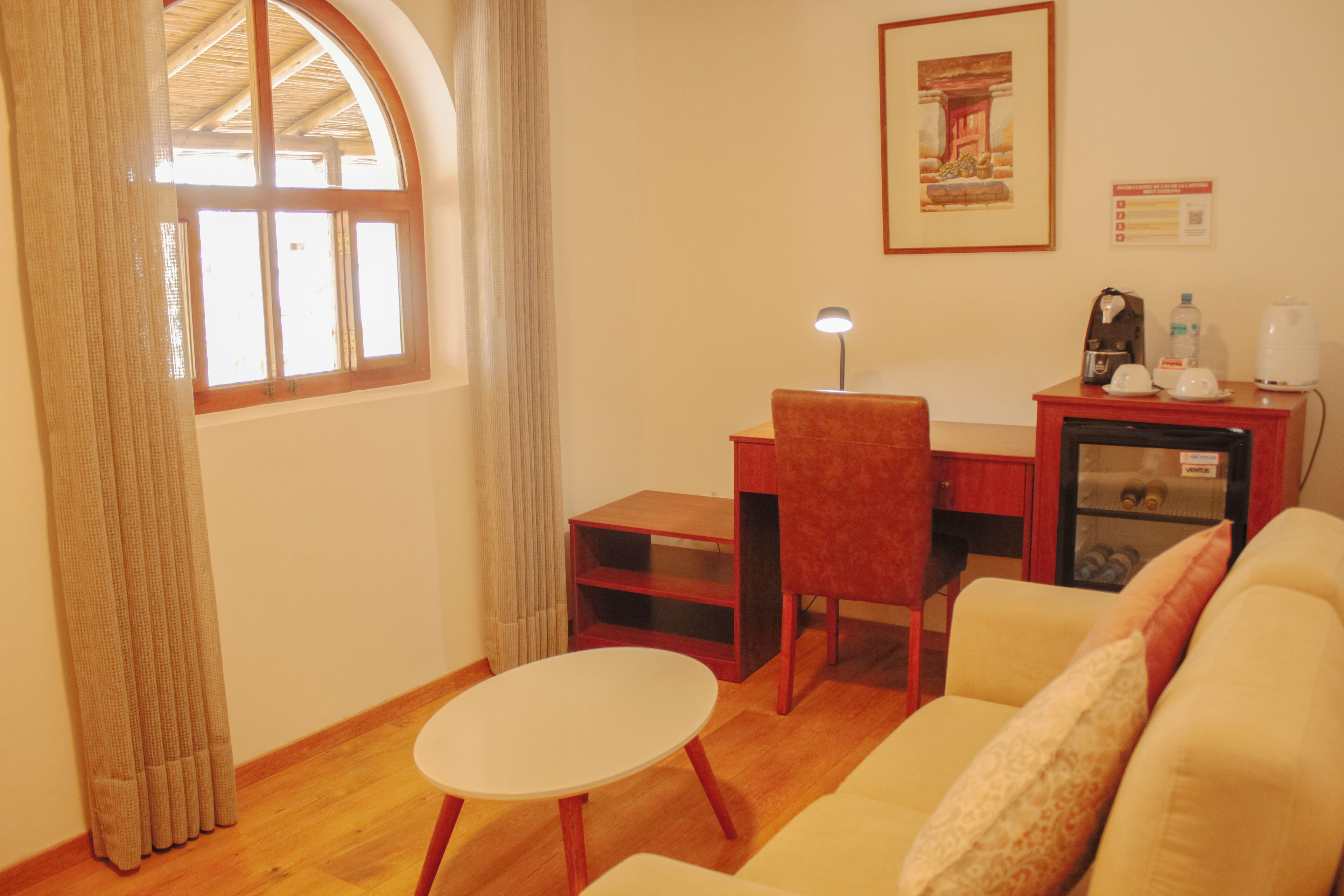 A room with a wooden floor, a white table, a red chair, a sofa, a desk with a lamp, a coffee maker, a water dispenser, and a framed picture on the wall.