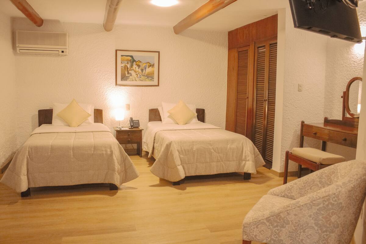 A bedroom with two beds, a wooden chair, a desk with a mirror, and a painting on the wall.