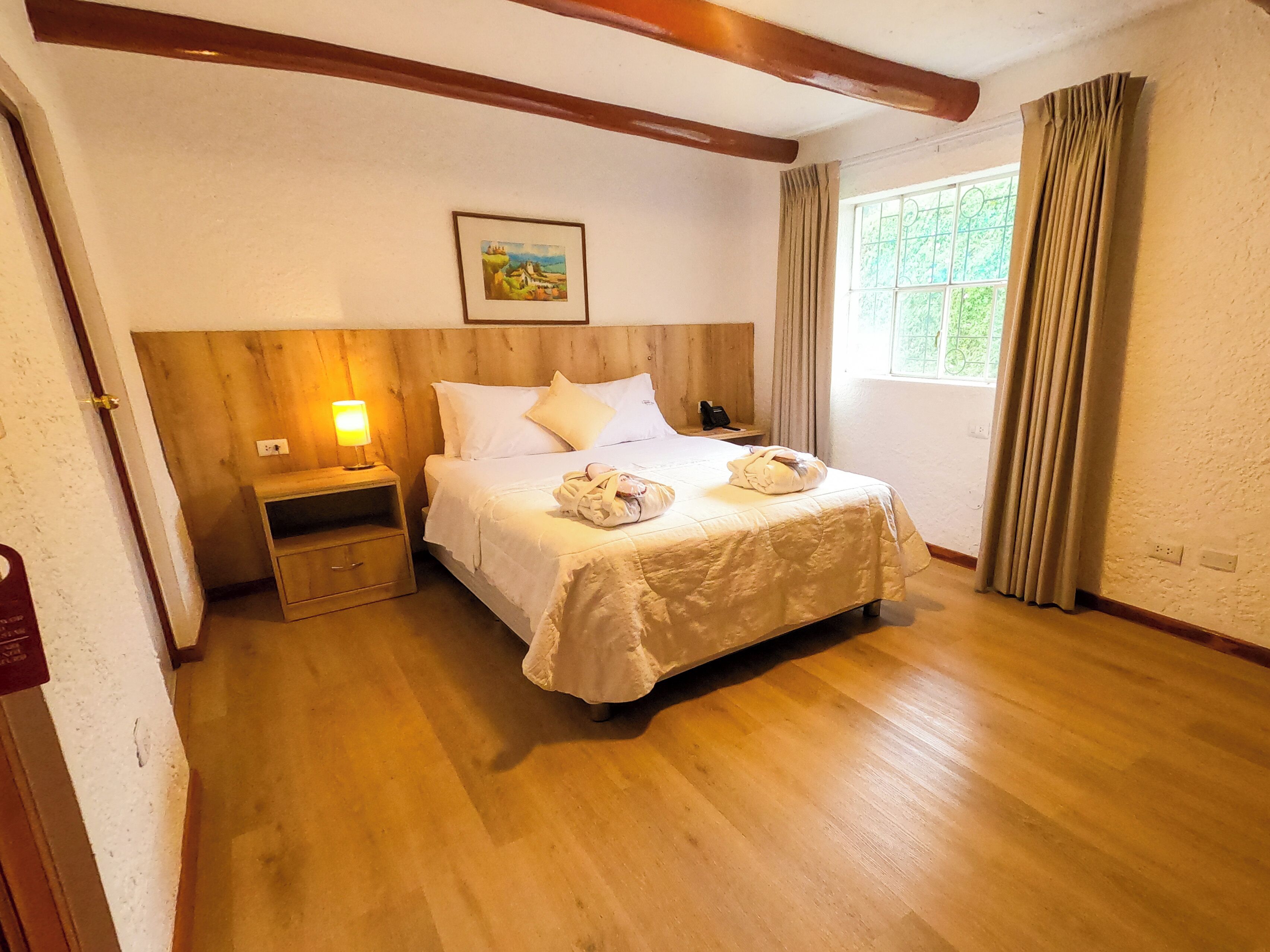 A neatly arranged bedroom with a wooden headboard, a bedside lamp, a framed picture on the wall, and a window with curtains.