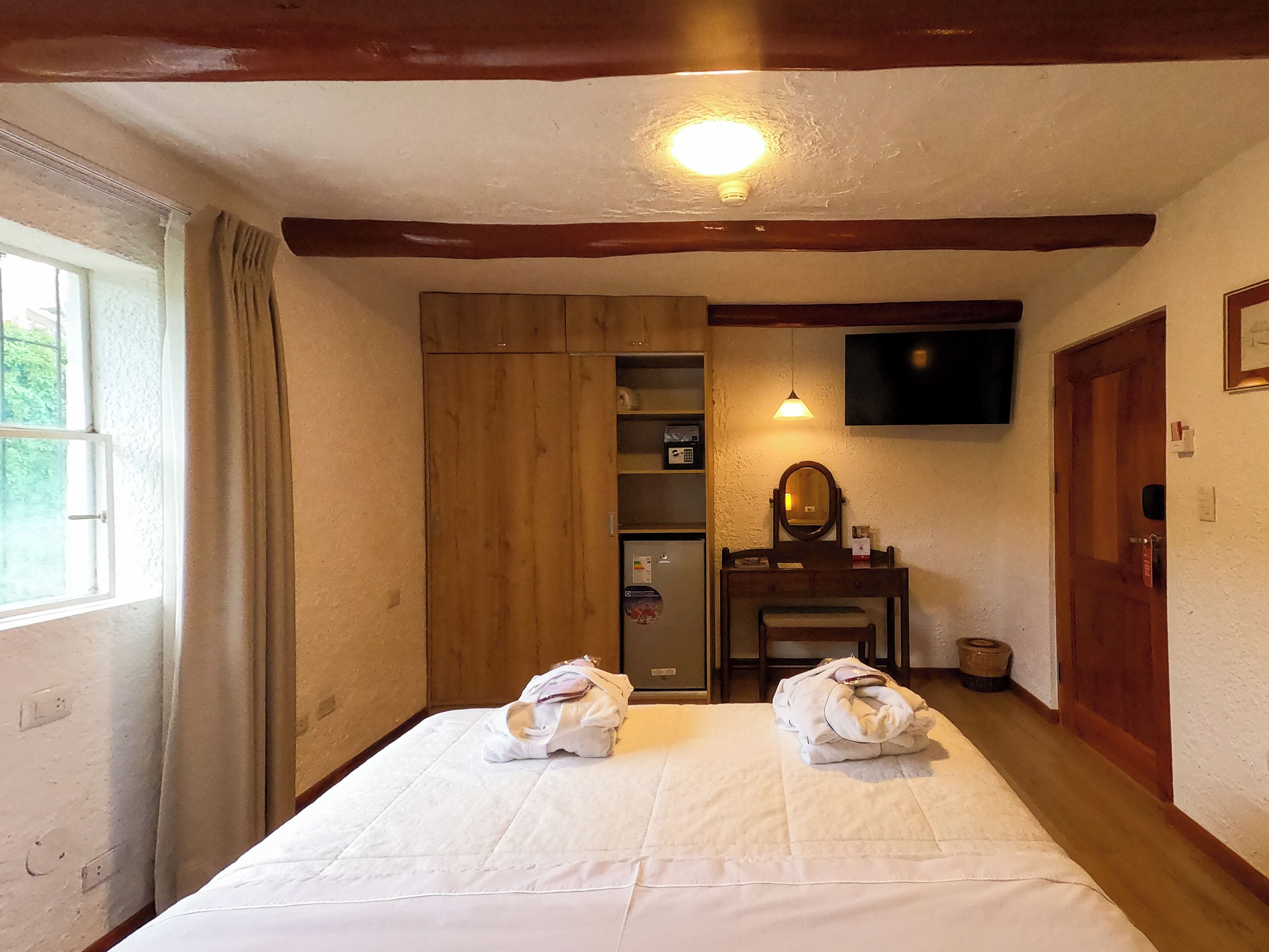 A hotel room with a bed, a wooden wardrobe, a television mounted on the wall, and a small table with a mirror.