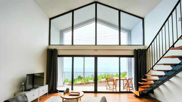 Een ruime woonkamer met een grote schuifpui naar een balkon met uitzicht op de oceaan. Aan de rechterkant een trap en aan de linkerkant een flatscreen-tv.